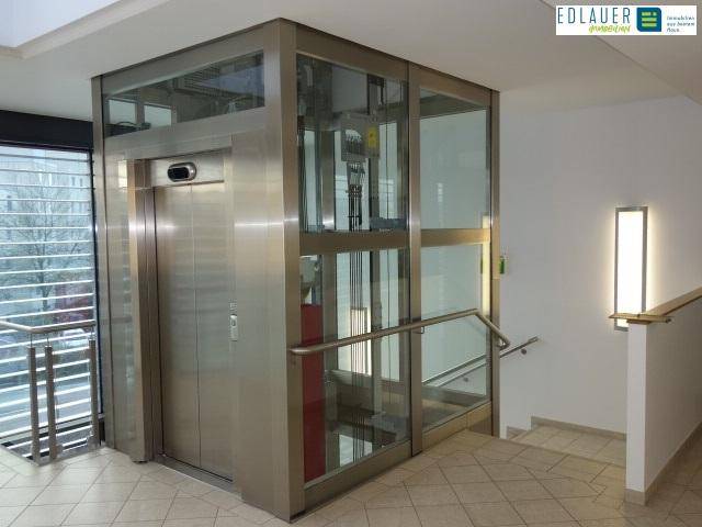 Moderner Glasaufzug in einem hellen Flur mit Fliesenboden und Handlauf, bietet barrierefreien Zugang.