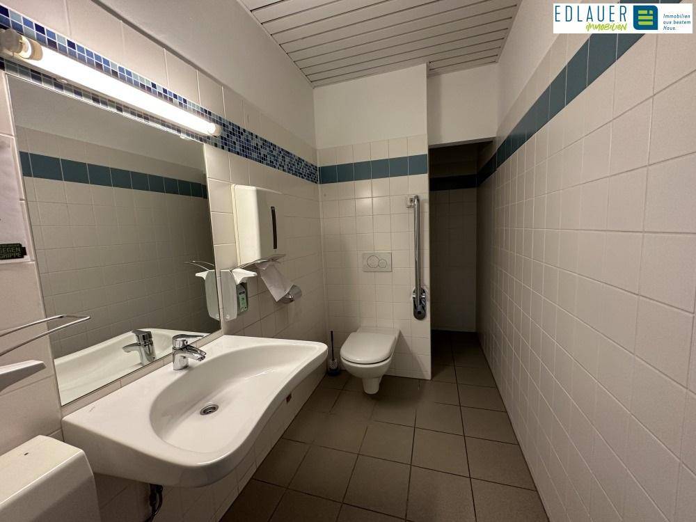 Sanitärraum mit weißen Fliesen, Waschbecken und Toilette, ausgestattet mit Haltegriffen für Barrierefreiheit.