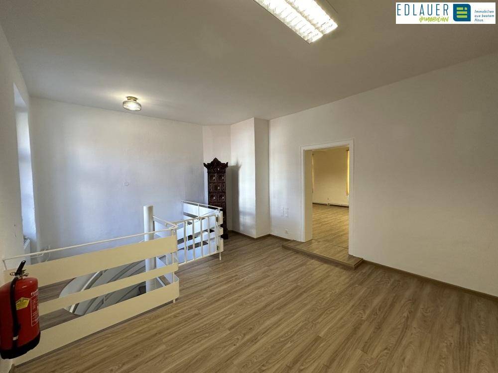 Heller Raum mit Laminatboden, Geländer und Kachelofen, der zu weiteren Räumen führt.