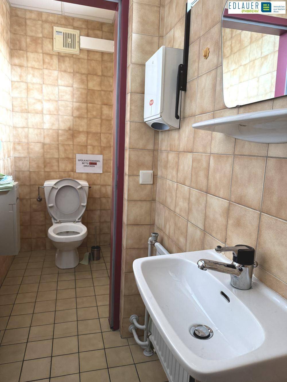 Toilette mit beigefarbenen Fliesen, Waschbecken und WC, ausgestattet mit einem Abluftventilator.