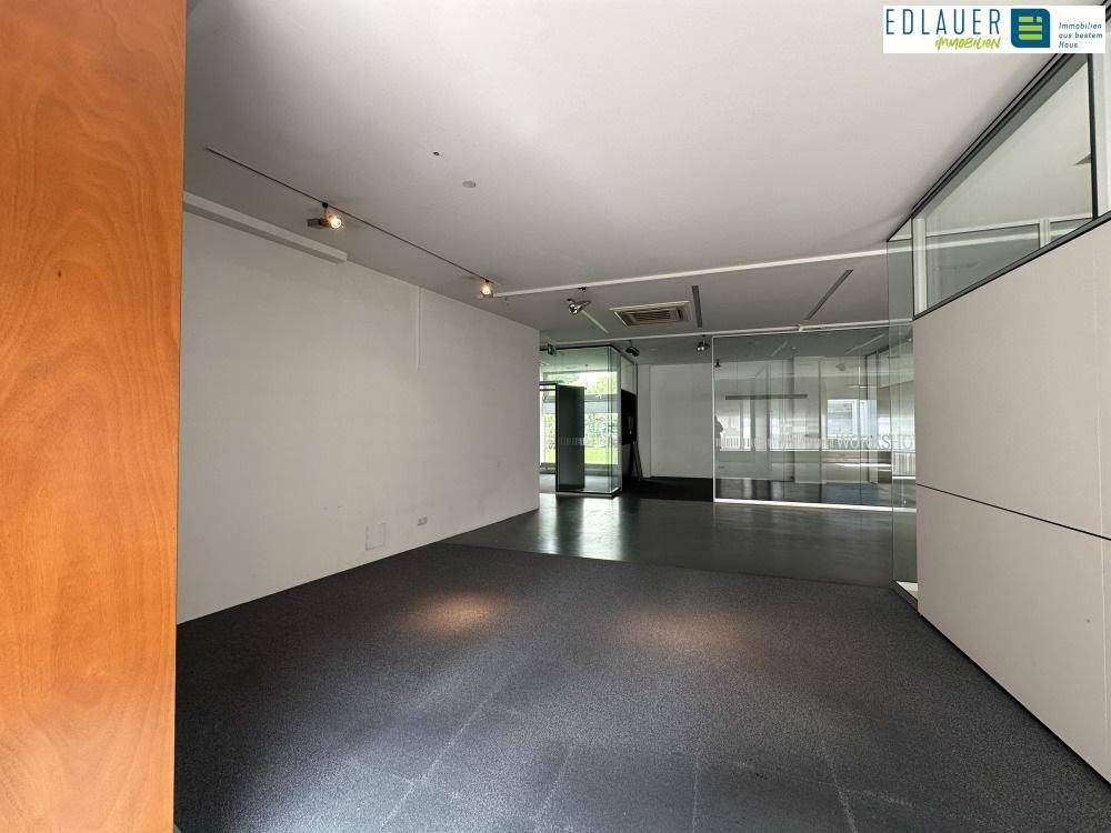 Geräumiger Büroraum mit dunklem Teppichboden, weißen Wänden und modernen Glas-Trennwänden, die Licht durchlassen.