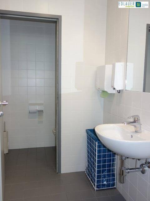 Helles Badezimmer mit Waschbecken, Spiegel und weißen Fliesen, das zu weiteren Sanitärbereichen führt.