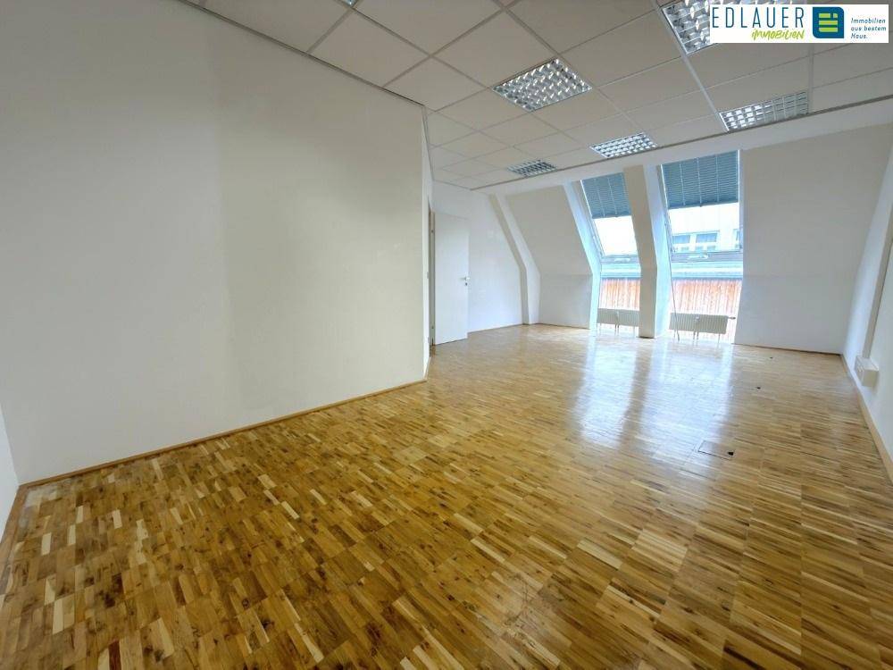Leerer Büroraum mit Holzparkettboden, weißen Wänden und schrägen Dachfenstern, die viel Licht hereinlassen.