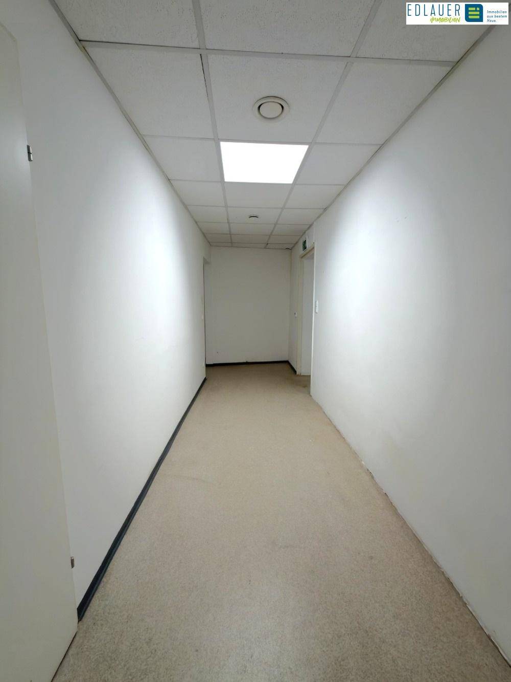 Langer, leerer Flur mit weißen Wänden, beigem Boden und einer Rasterdecke mit integrierter Beleuchtung.