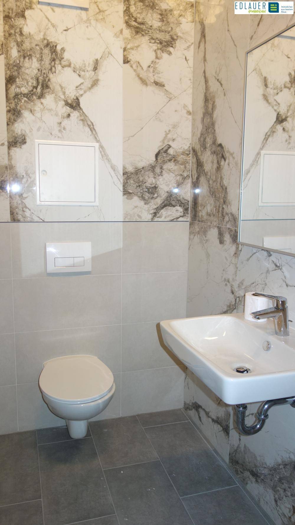 Modernes Badezimmer mit Waschbecken, Spiegel, Wand-WC und Fliesen in Marmor-Optik.