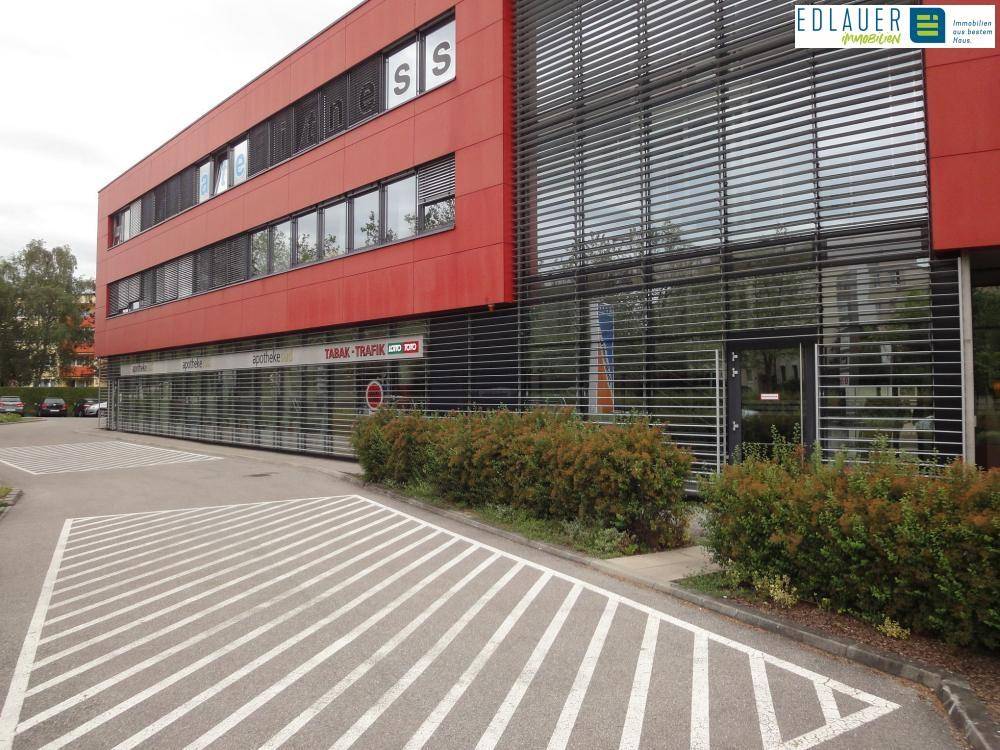 Rotes Geschäftsgebäude mit großen Fenstern und Parkplätzen davor, beherbergt verschiedene Geschäfte.