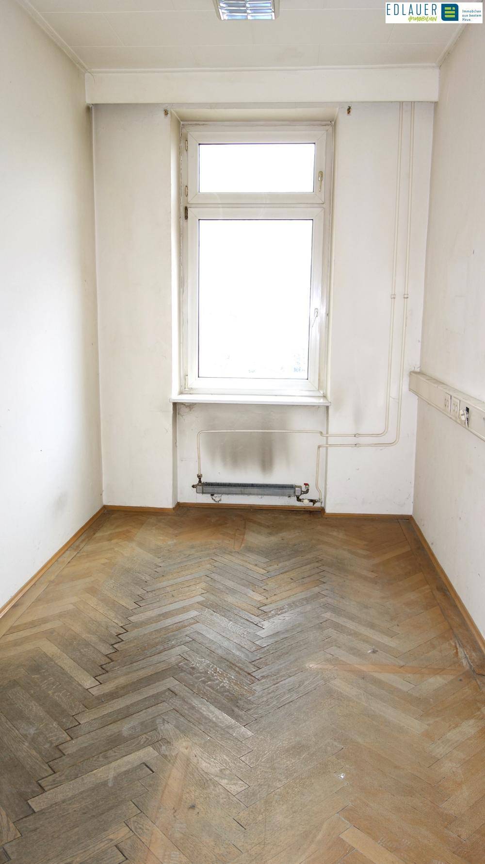 Schmaler Raum mit Fischgrätparkett, einem Fenster und einem Heizkörper unter der Fensterbank.