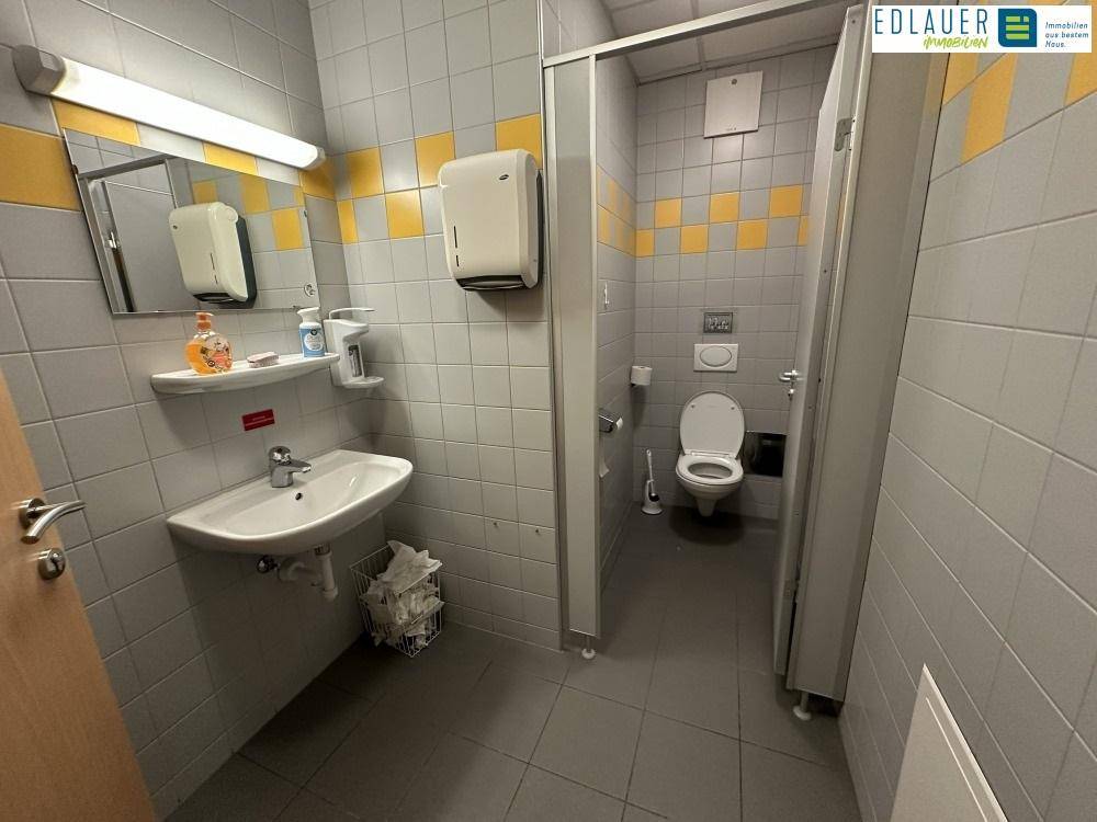 Öffentliche Toilette mit mehreren Kabinen, Waschbecken und Fliesen in Grau und Gelb, funktional und sauber.