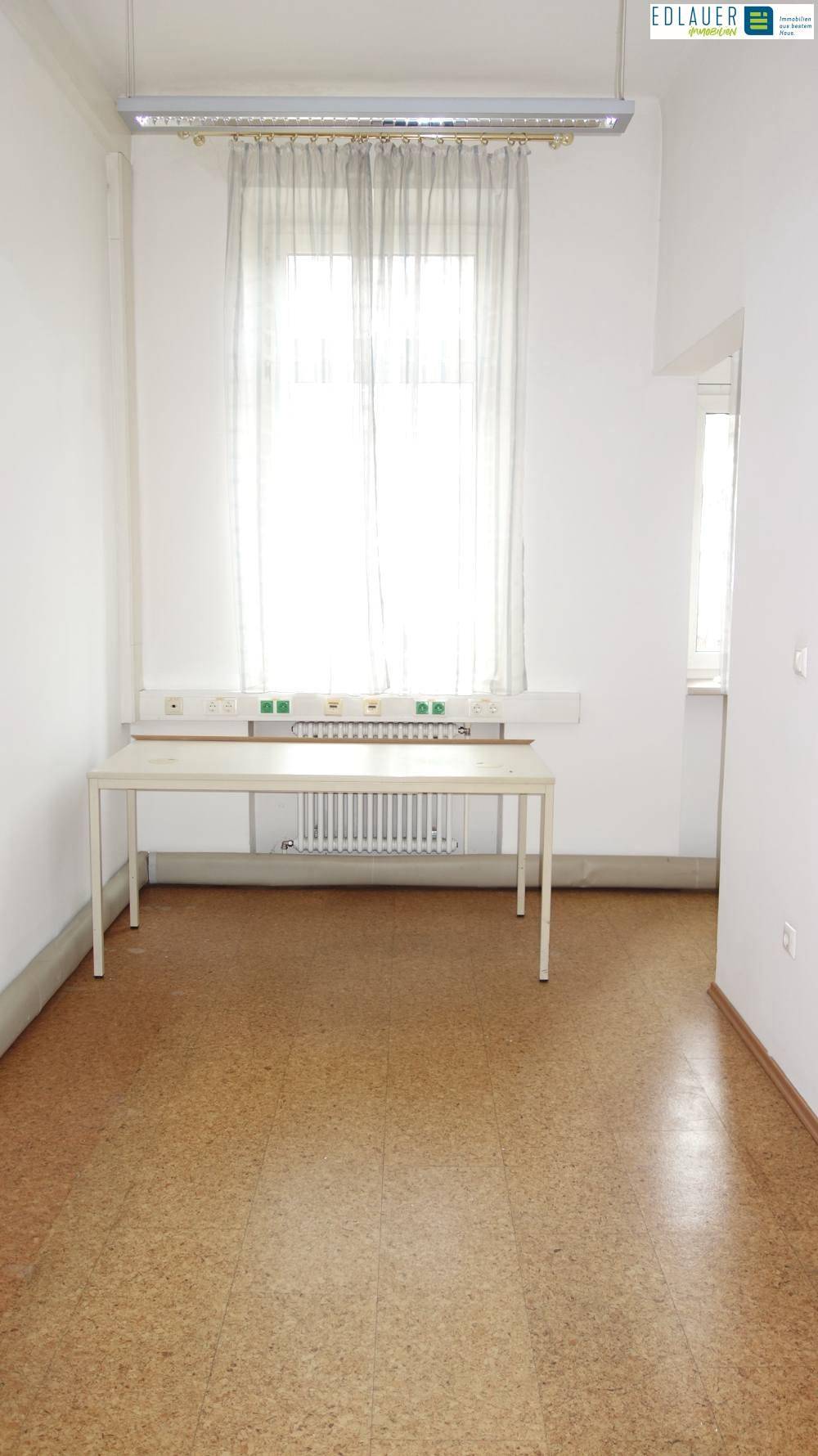 Einzelner Büroraum mit einem Schreibtisch unter einem großen Fenster, ausgestattet mit Linoleumboden und Deckenleuchte.
