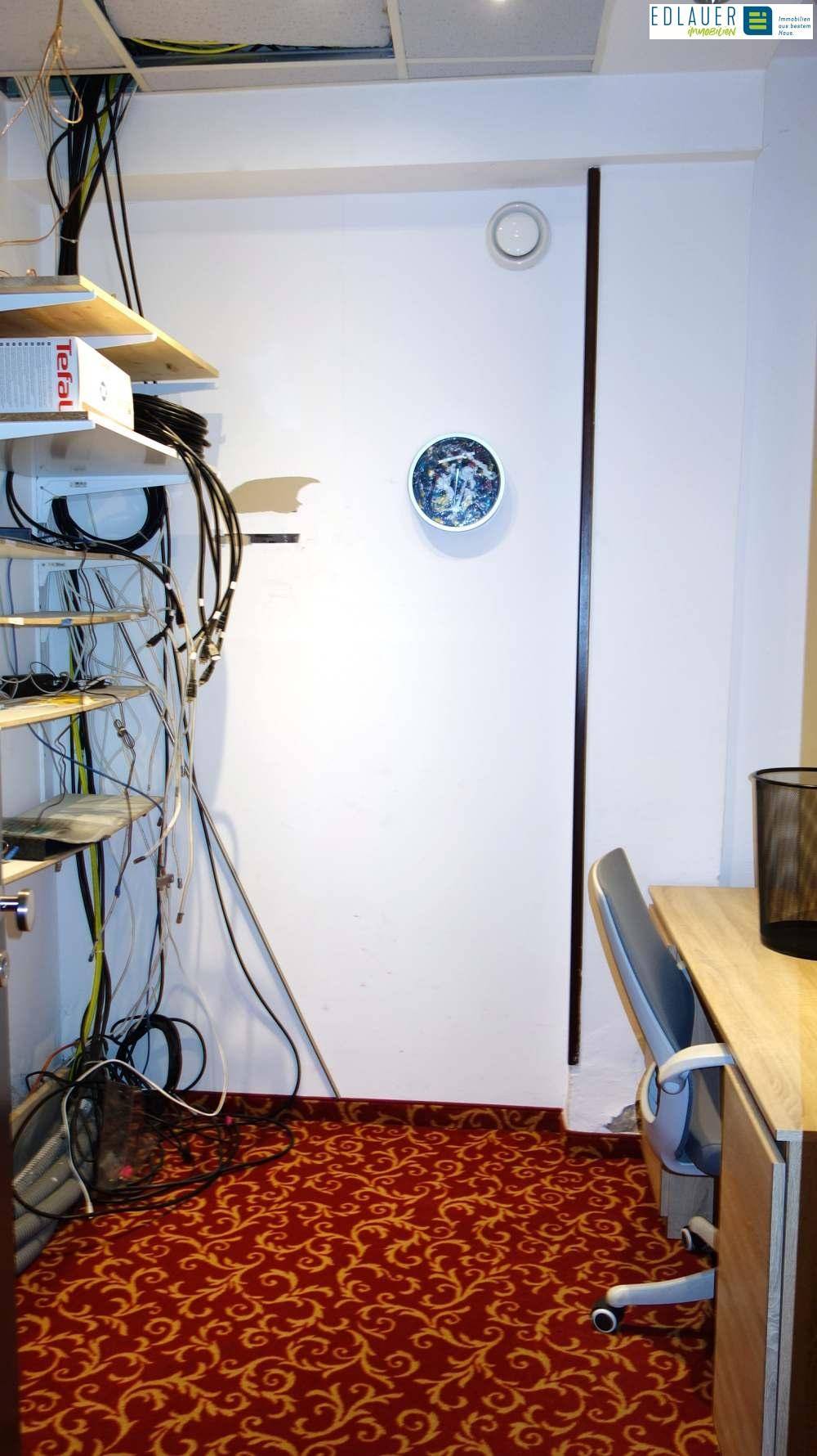 Raum mit unordentlichen Kabeln, einem Schreibtisch und rotem gemustertem Teppichboden, möglicherweise ein Serverraum oder Technikraum.