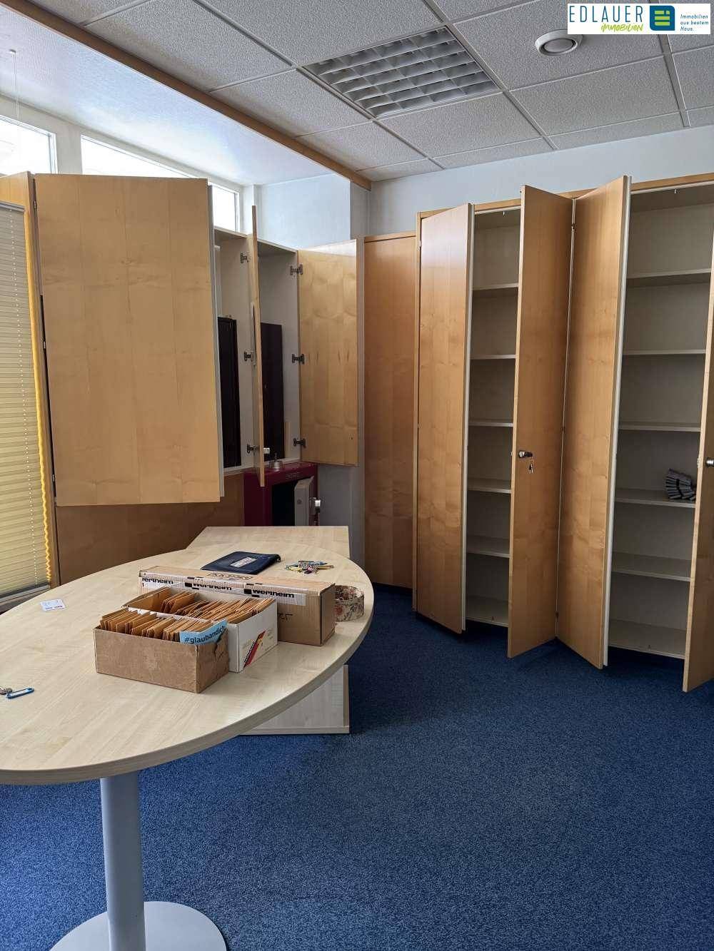 Büroraum mit blauem Teppichboden, Schreibtisch und offenen Holzschränken für Ablage.