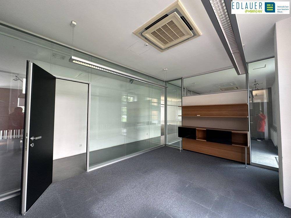 Moderner Büroraum mit Glastüren, Teppichboden und einem eleganten Holzregal als Raumteiler.