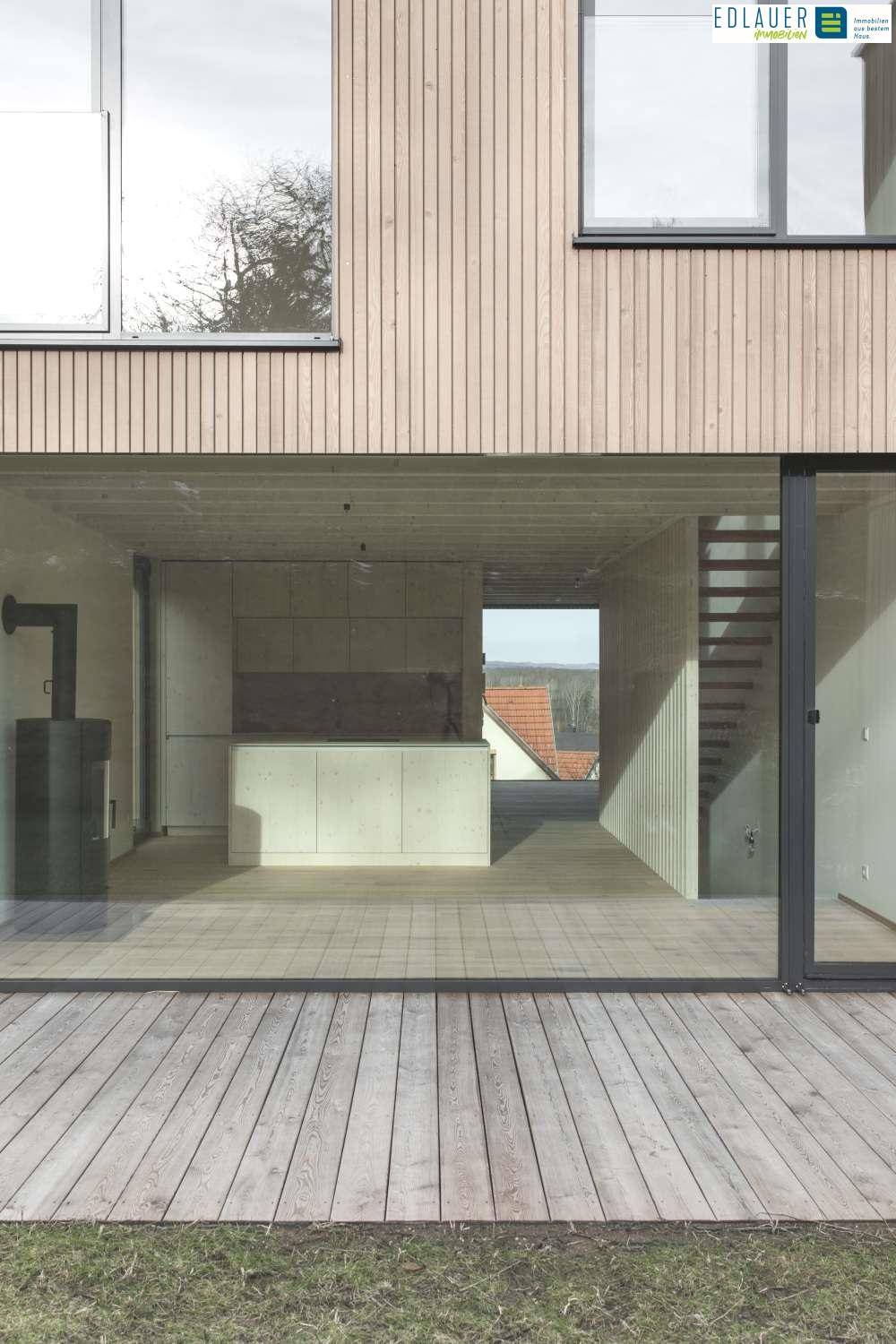 Heller Wohnbereich mit Holzboden, Kaminofen und Zugang zu einer Holzterrasse durch große Glasfronten.