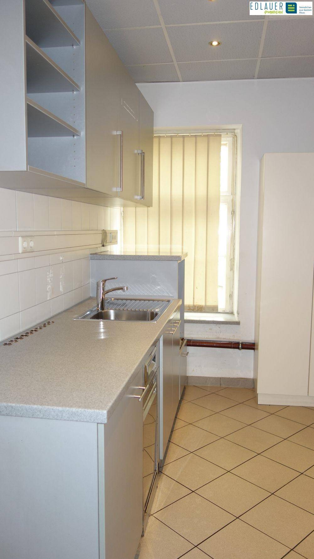 Kleine Küchenzeile mit grauen Schränken, Spüle und einem Fenster mit Jalousien, ideal für eine Büroküche.