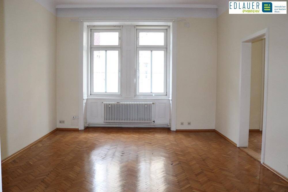 Geräumiges Zimmer mit klassischem Fischgrätparkett und großen Fenstern, die viel Tageslicht hereinlassen.