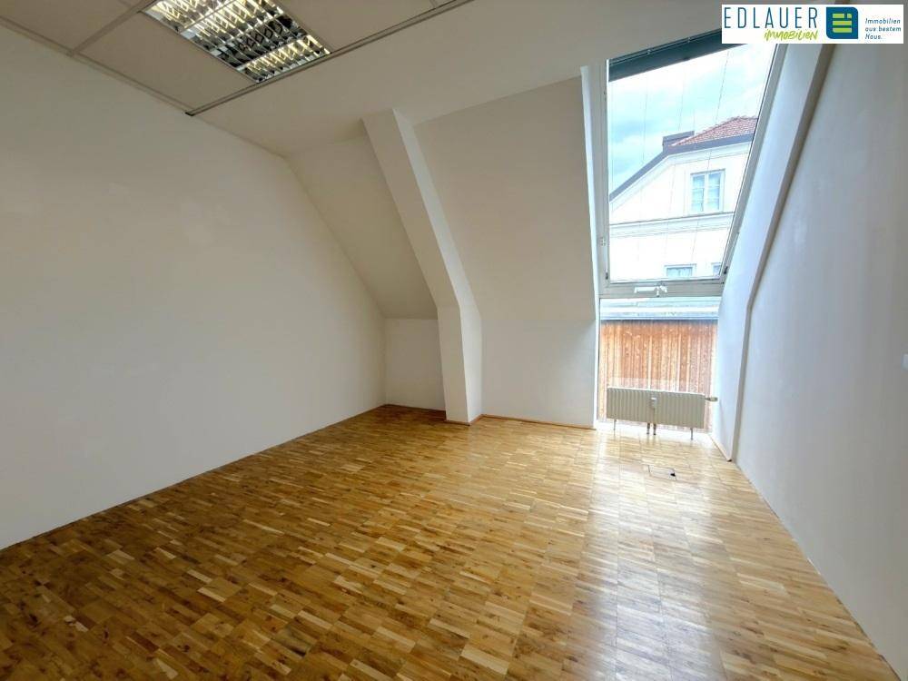 Leerer Büroraum mit Holzparkett, weißen Wänden und einem großen, schrägen Fenster, das Tageslicht spendet.