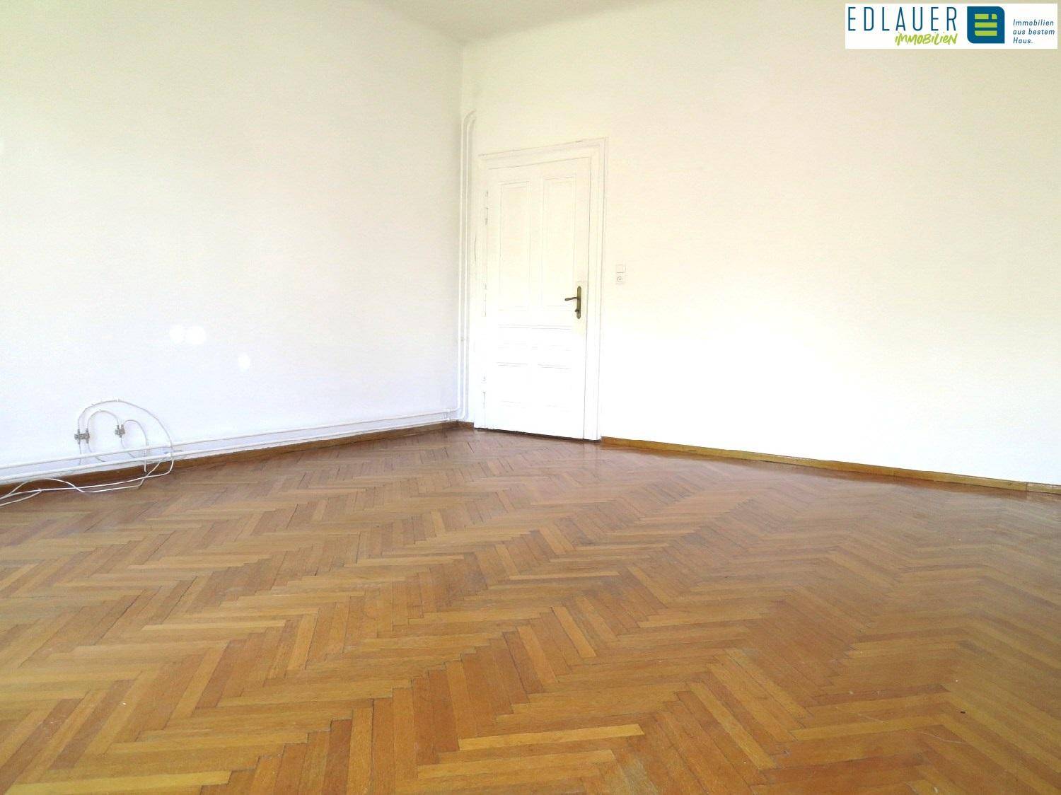 Geräumiges Zimmer mit klassischem Fischgrätparkett und einer weißen Holztür.
