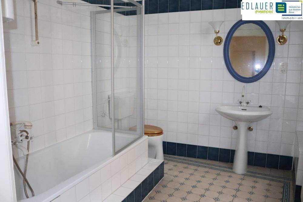 Gefliestes Badezimmer mit Badewanne, Duschabtrennung, Waschbecken und ovalem Spiegel.