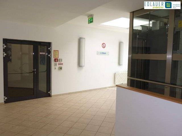 Heller Flur mit Fliesenboden und modernem Glasaufzug, führt zu weiteren Räumen im Gebäude.