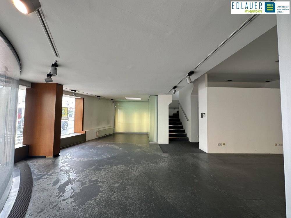 Geräumiger Ladenbereich mit dunklem Boden, einer Treppe und großen Schaufenstern zur Straße, die viel Licht hereinlassen.