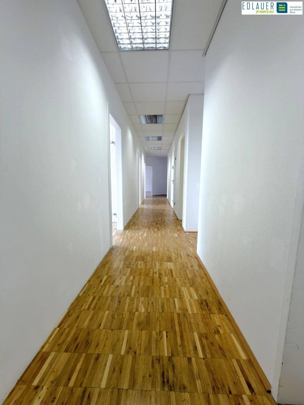Langer Flur mit Holzparkettboden, weißen Wänden und Deckenbeleuchtung, der zu mehreren Räumen führt.