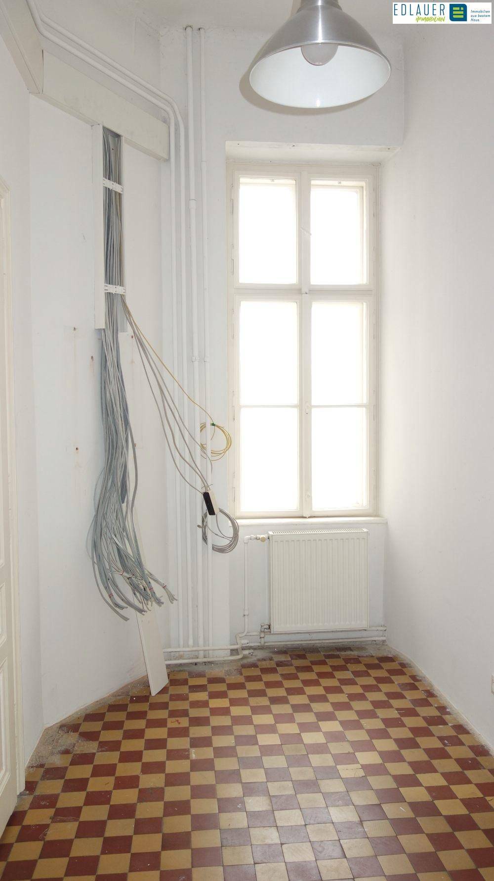 Raum mit kariertem Fliesenboden, einem Fenster und sichtbaren Kabeln, möglicherweise ein Abstellraum.