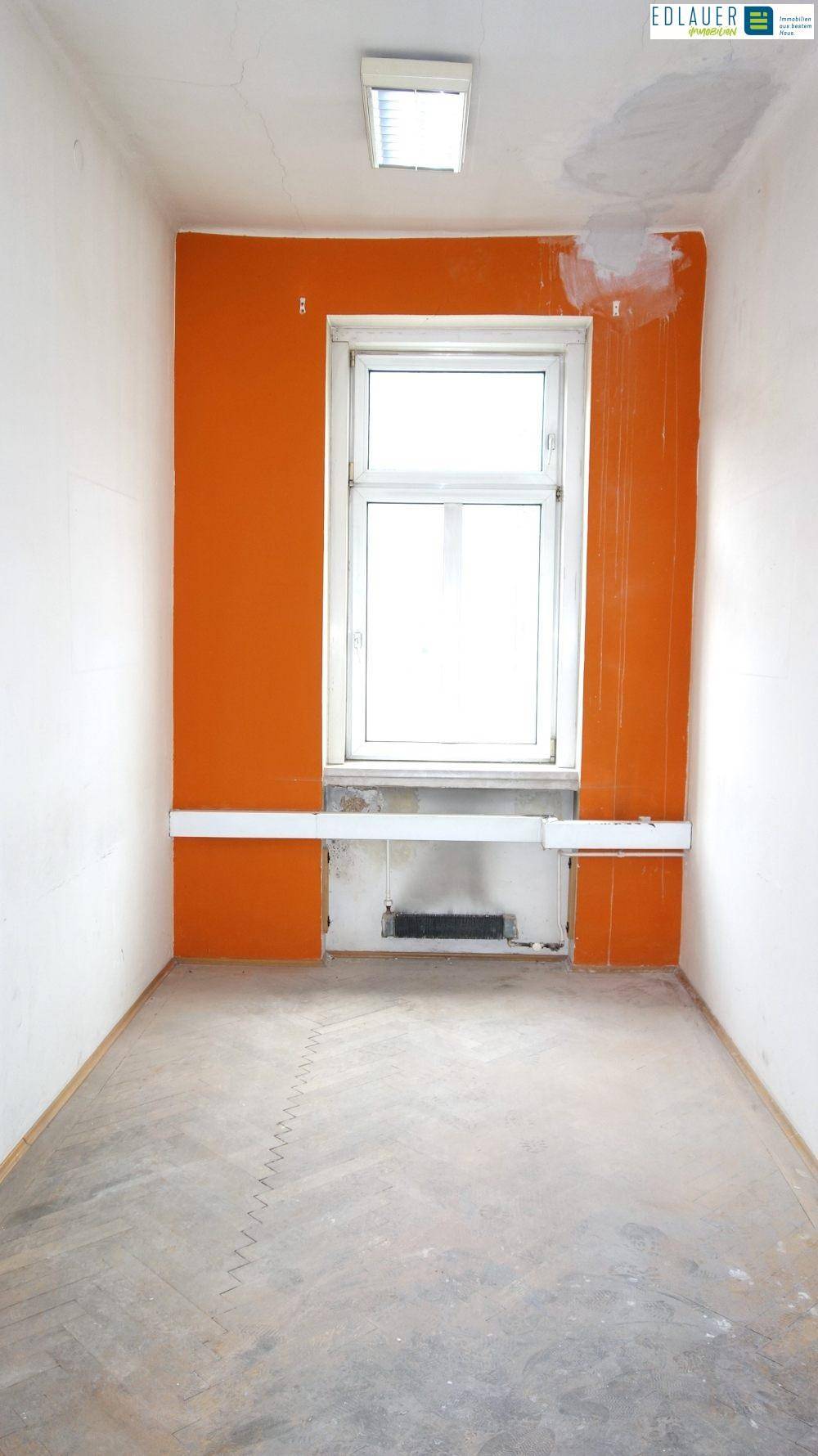 Schmaler Raum mit orangefarbener Akzentwand, einem Fenster und einem Betonboden.