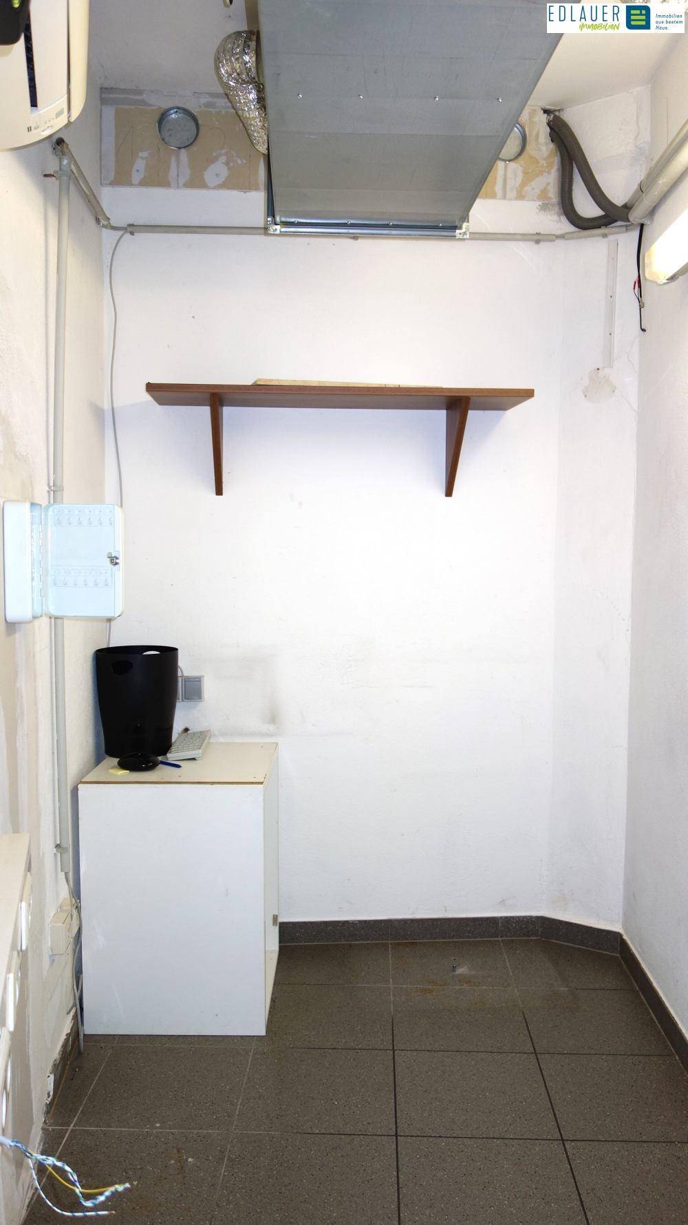 Kleiner Abstellraum mit Fliesenboden, einem Regal und sichtbaren Lüftungsrohren an der Decke für technische Installationen.