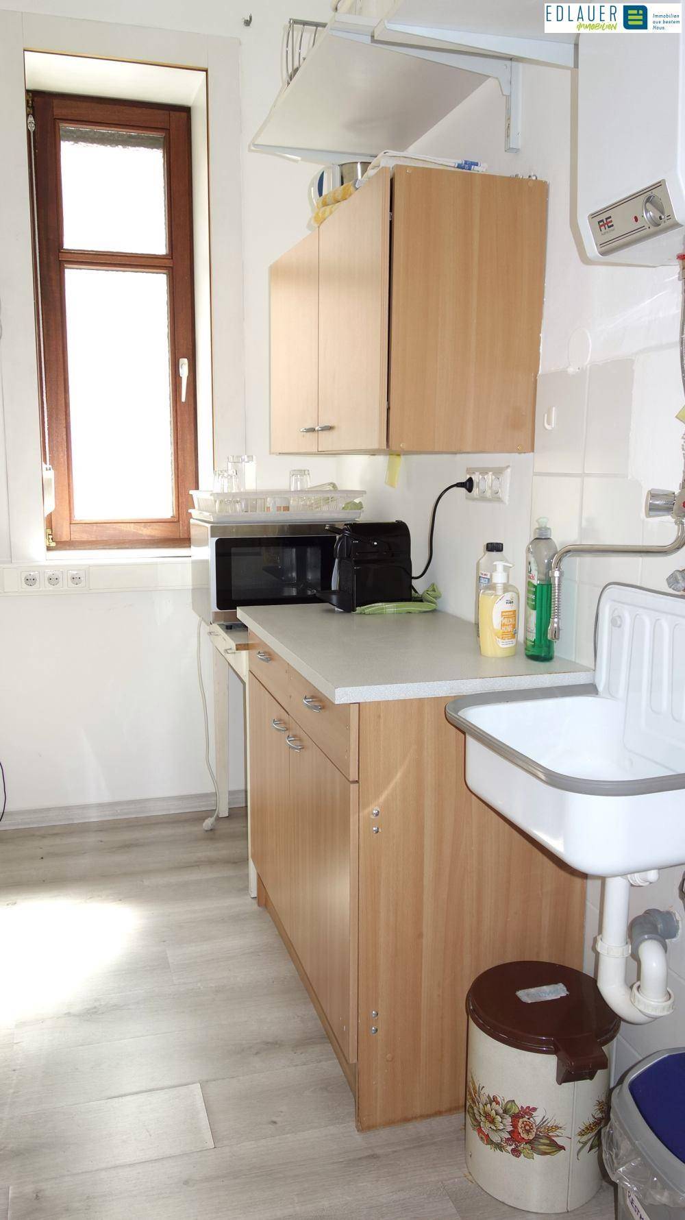 Kleine Küchenzeile mit Spüle, Mikrowelle und Holzschränken, gut beleuchtet durch ein Fenster.