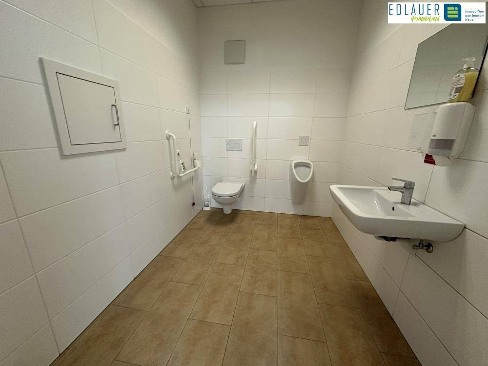 Barrierefreie Toilette mit Haltegriffen, Urinal und Waschbecken, hell gefliest und geräumig.