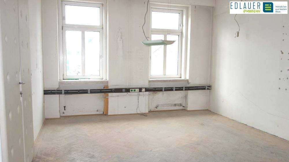 Renovierungsbedürftiger Raum mit zwei Fenstern, freiliegenden Kabeln und Betonboden.