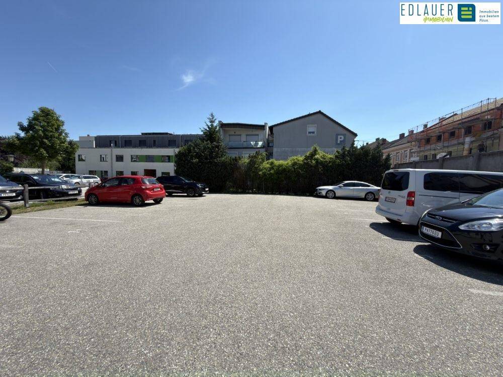 Weitläufiger Parkplatz im Freien mit geparkten Autos und einer grünen Hecke im Hintergrund unter klarem Himmel.