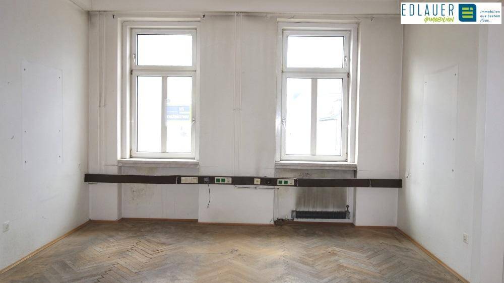 Heller Raum mit zwei Fenstern, Fischgrätparkett und einer langen Installationsleiste an der Wand.