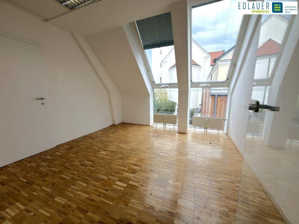 Heller Büroraum mit Holzparkettboden und großen, schrägen Fenstern, die einen Blick ins Grüne bieten.