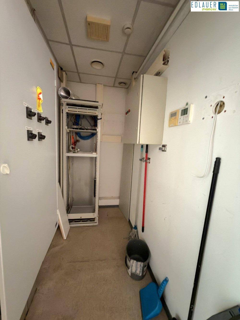 Technikraum mit Serverrack, Warmwasserbereiter und Reinigungsutensilien, zeigt funktionale Ausstattung.