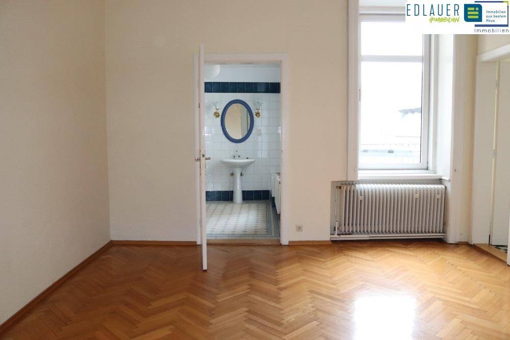 Heller Wohnraum mit Fischgrätparkett und Blick in ein gefliestes Badezimmer mit Fenster.