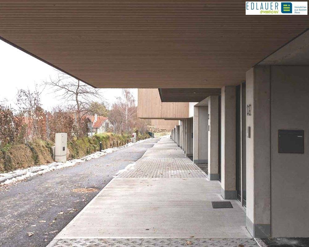 Überdachter Bereich unter den Häusern mit Betonweg und Parkplätzen, umgeben von kahlen Bäumen.