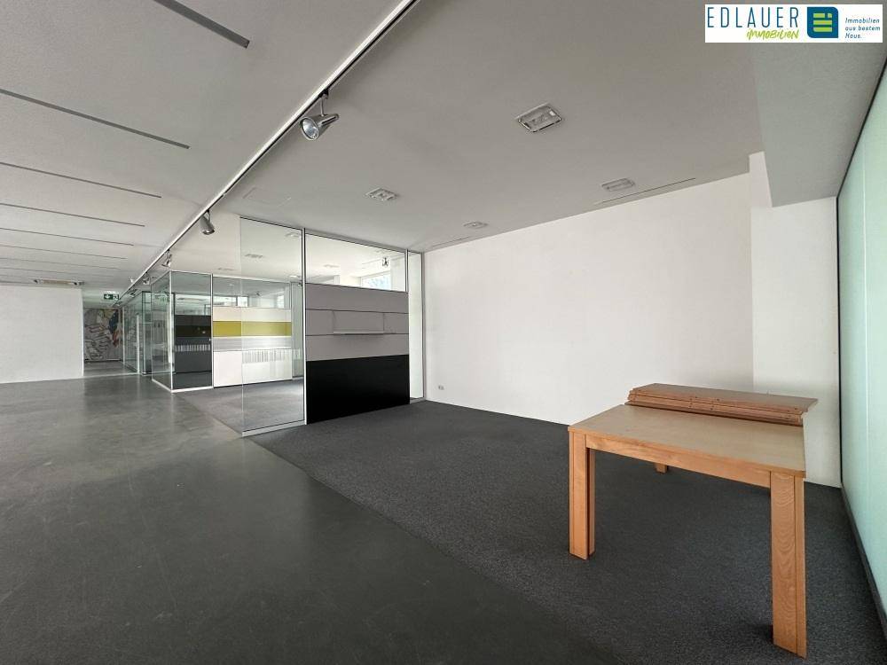 Offener Bürobereich mit Teppichboden, einem Holztisch und modernen Glaswänden, die eine flexible Nutzung ermöglichen.
