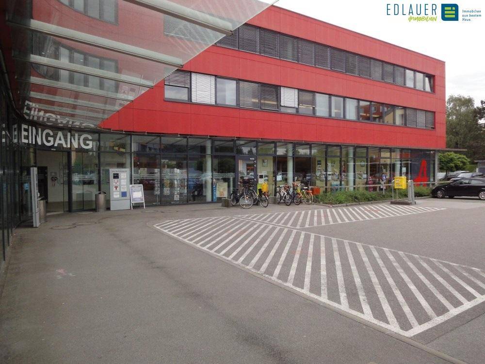 Modernes rotes Gebäude mit großem Eingangsbereich und Fahrradstellplätzen, umgeben von Parkflächen.