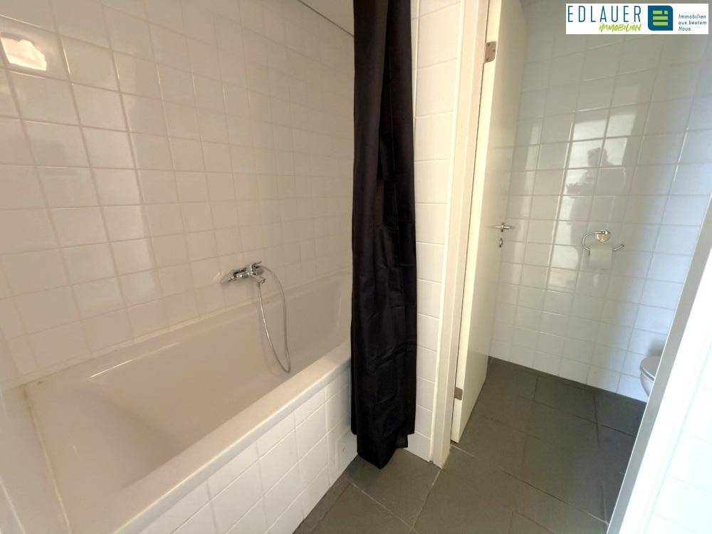 Badezimmer mit Badewanne, weißen Wandfliesen und dunklem Boden, sowie einem schwarzen Duschvorhang.