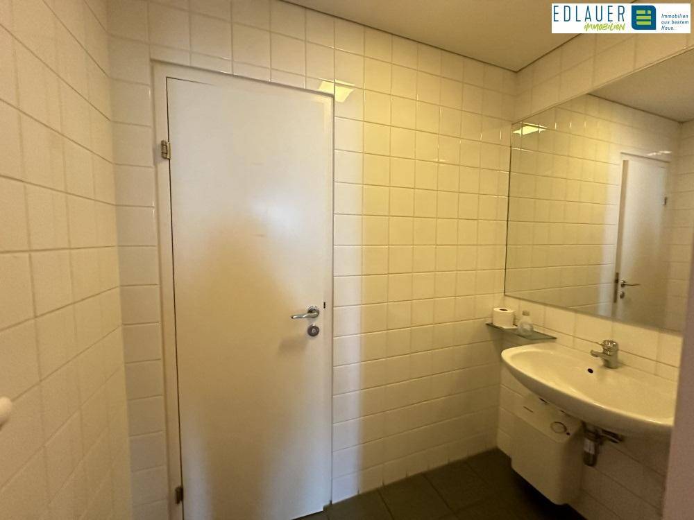 Badezimmer mit weißen Fliesen, einem Waschbecken, Spiegel und einer Tür, in einem gepflegten Zustand.