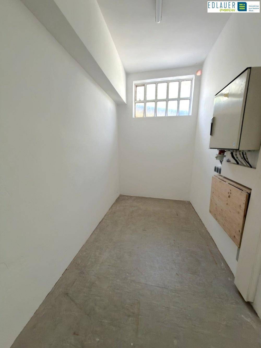 Schmaler Lagerraum mit Betonboden, weißen Wänden und einem hochgelegenen Fenster für Tageslicht.