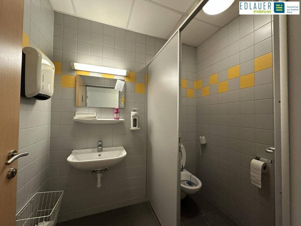 Sanitärbereich mit Waschbecken, Spiegel und Toilettenkabinen, gefliest in Grau und Gelb, gut gepflegt.
