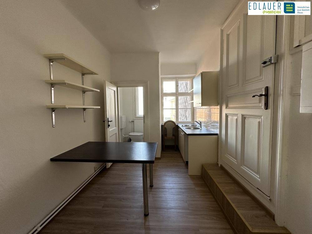 Kompakte Küchenzeile mit Spüle, Schränken und Fenster, ergänzt durch einen Klapptisch und Regale. Eine Tür führt zu einem Badezimmer.