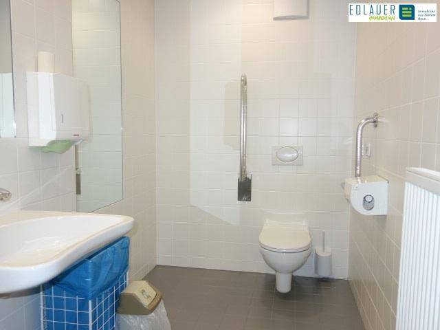 Barrierefreies Badezimmer mit weißen Fliesen, Toilette, Waschbecken und Haltegriffen für zusätzliche Sicherheit.