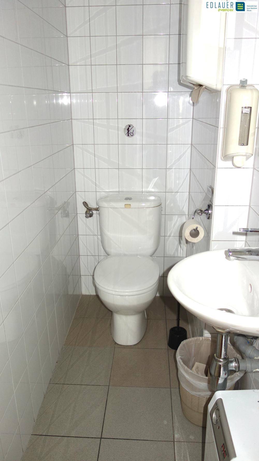 Einfache Toilette mit weißen Fliesen an Wänden und Boden, ausgestattet mit WC, Waschbecken und Spender.
