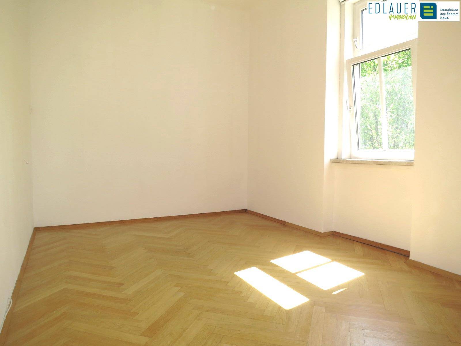 Sonniger Raum mit Fischgrätparkett und einem großen Fenster, das viel Tageslicht hereinlässt.
