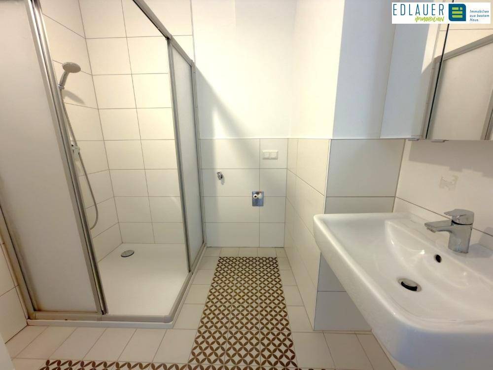 Helles Badezimmer mit Duschkabine und Waschbecken, ergänzt durch einen Spiegel und weiße Wandfliesen.