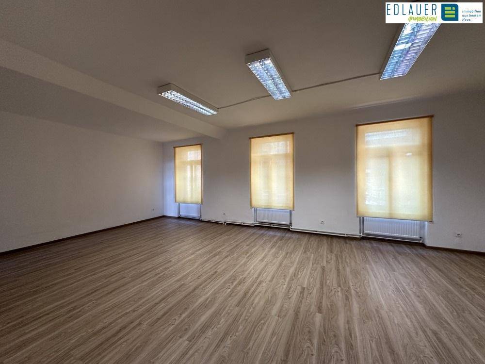 Heller Gewerberaum mit Laminatboden, drei Fenstern mit Jalousien und Heizkörpern, gut belichtet.