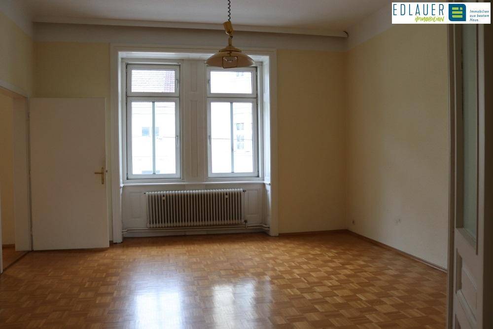 Helles Zimmer mit Fischgrätparkett, großen Fenstern und einem Heizkörper unter der Fensterbank.